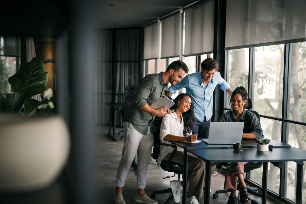 Vier Personen arbeiten lächelnd an einem Laptop in einem modernen Büro mit großen Fenstern.