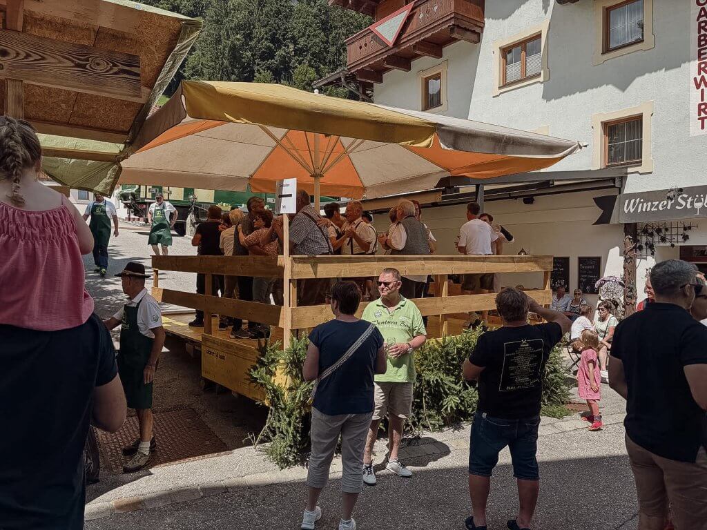 Menschen auf einer Terrasse unter Sonnenschirmen bei einem Fest vor einem Gebäude mit der Aufschrift 'Winzer'. Hintergrund mit Bäumen.