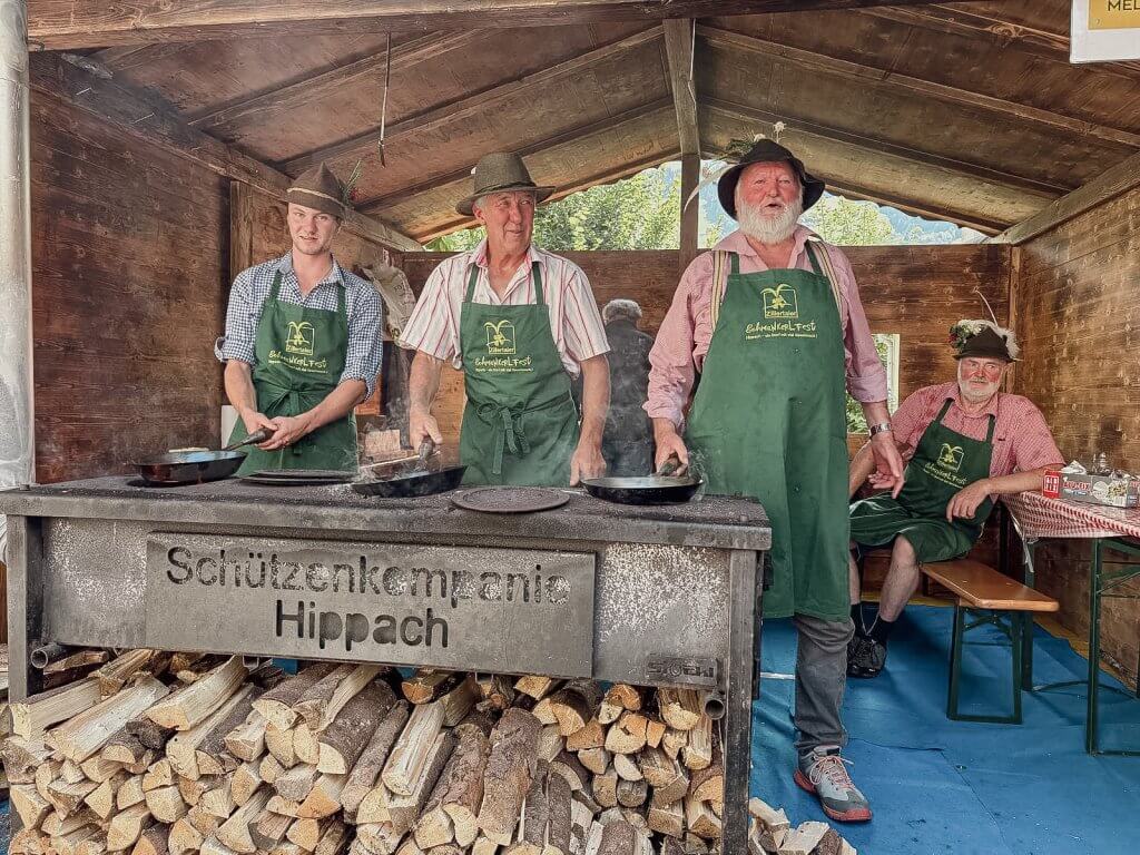 Vier Männer in traditionellen Trachten und Schürzen stehen an einem Holzofen und braten in einer Holzhütte.