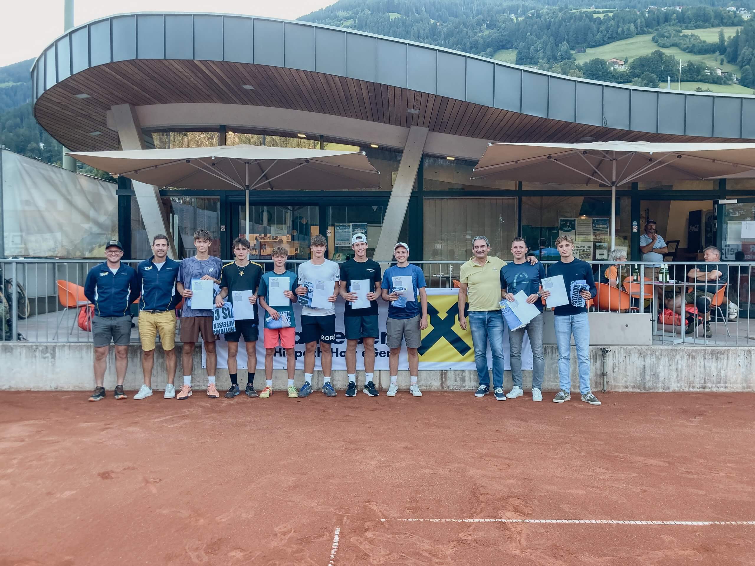 Gruppe von Männern mit Urkunden auf Tennisplatz vor modernem Gebäude.