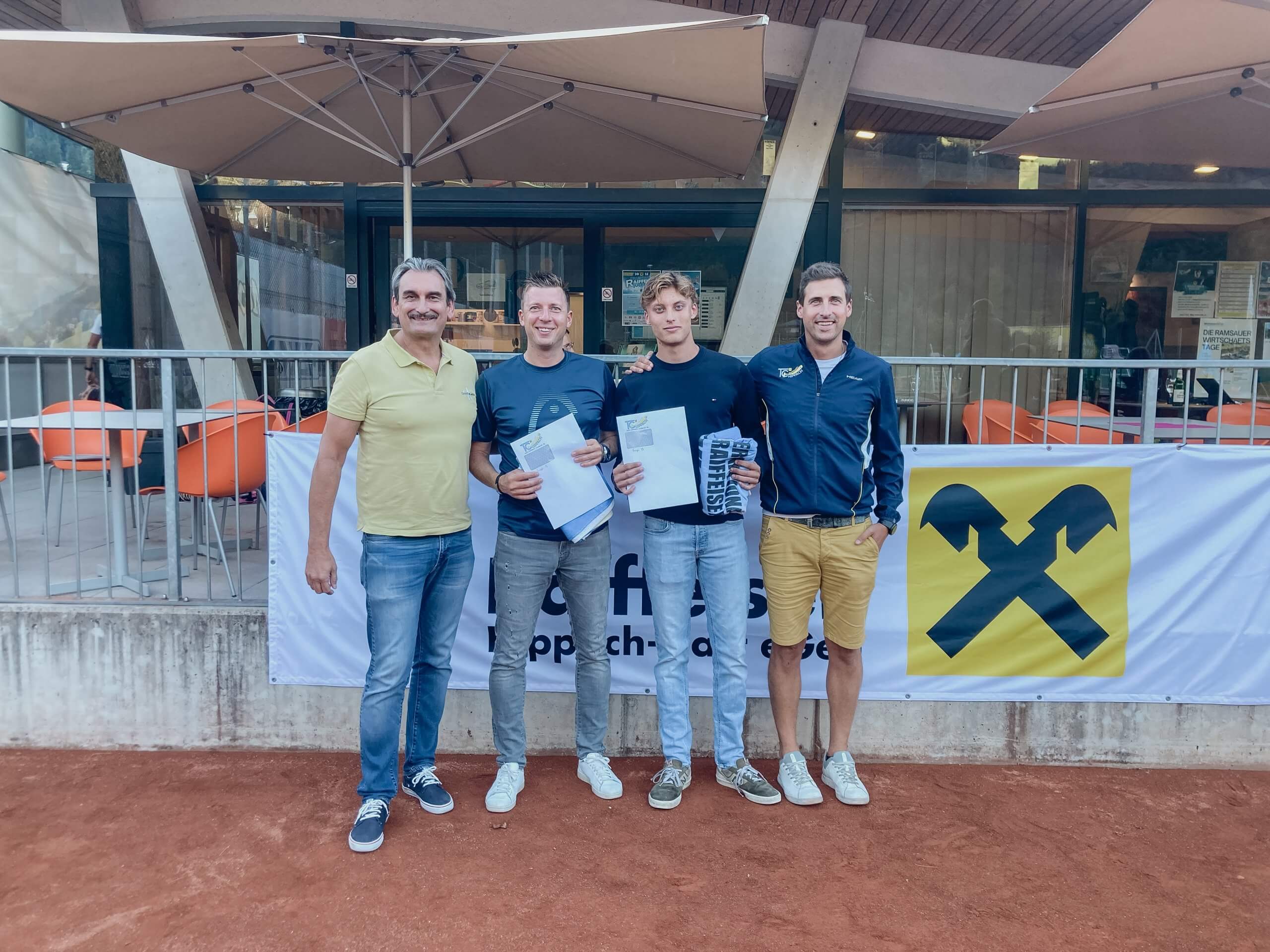 Vier Männer stehen vor einem Gebäude auf einem Tennisplatz, zwei halten Zertifikate, im Hintergrund ein Raiffeisen-Banner.