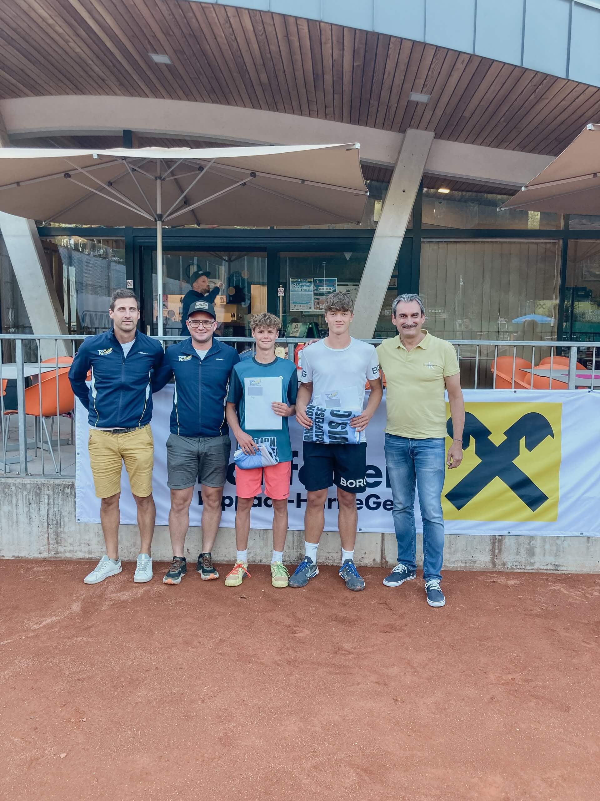 Fünf Männer posieren auf einem Tennisplatz vor einem Gebäude mit Sonnenschirmen und einem Werbebanner.