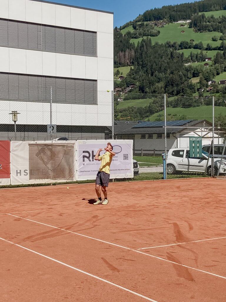 Tennisspieler auf rotem Sandplatz vor einer ländlichen Kulisse mit Bergen und Gebäuden im Hintergrund.