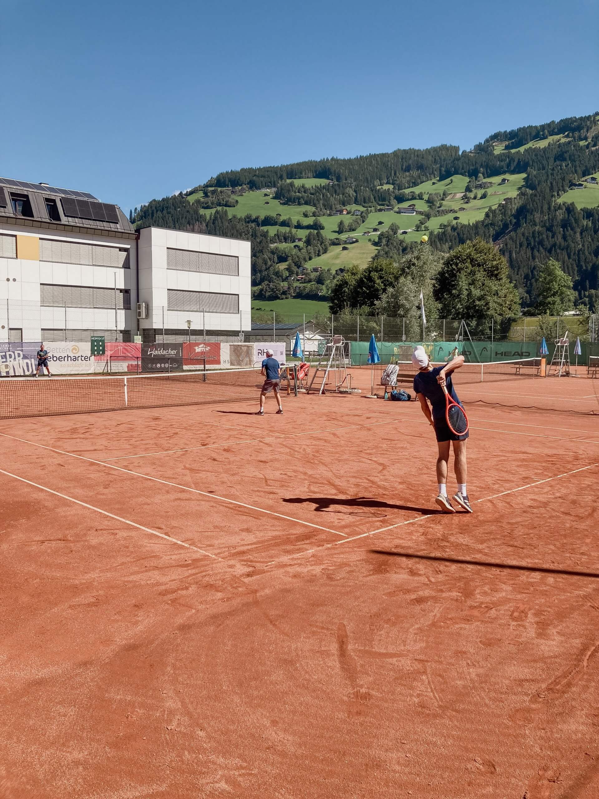 Tennisspiel auf rotem Sandplatz mit Bergen im Hintergrund.