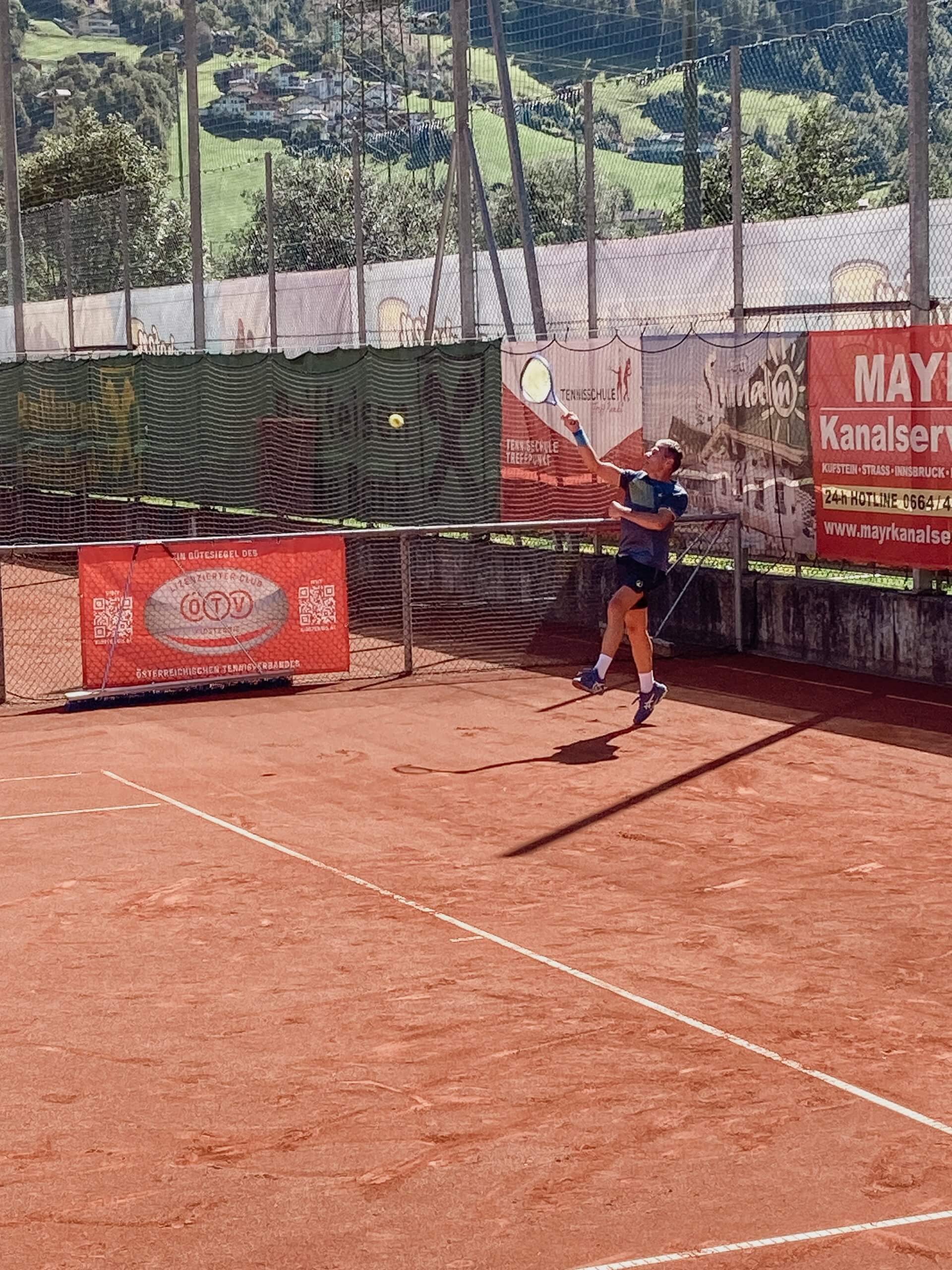 Tennisspieler schlägt auf Sandplatz einen Ball, umgeben von Werbebannern, mit grünen Hügeln im Hintergrund.