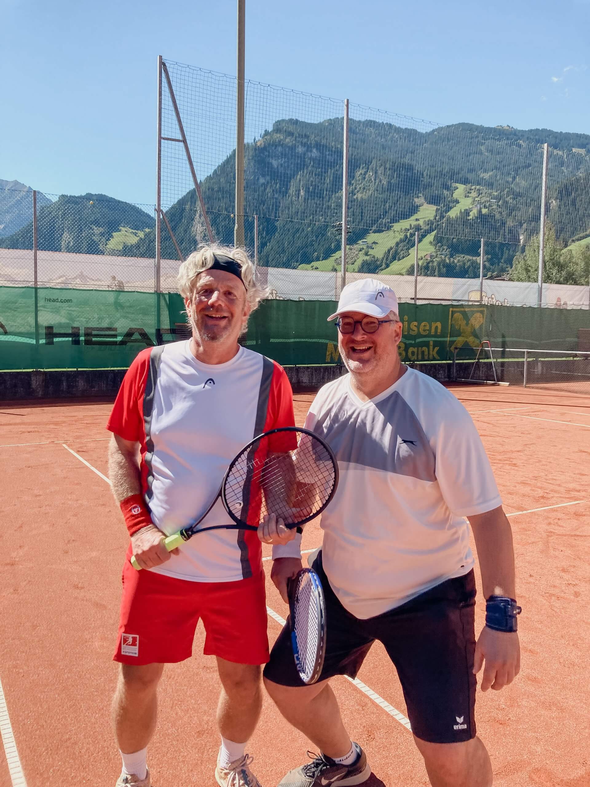 Zwei Männer in Sportkleidung mit Tennisschlägern auf rotem Tennisplatz, Berge im Hintergrund.