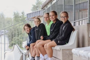Fünf Mädchen in Sportkleidung sitzen lächelnd auf Tribünenstühlen in einem Stadion.