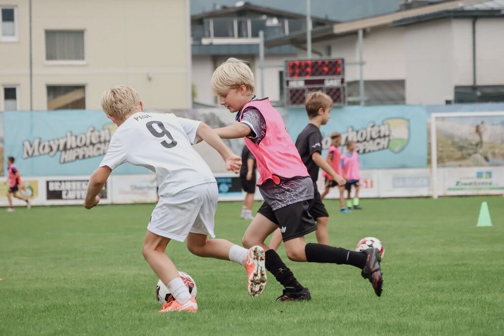 Kinder spielen auf einem Fußballfeld, zwei Jungen im Vordergrund kämpfen um den Ball.