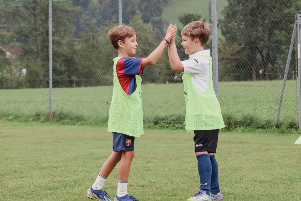 Zwei Jungen auf einem Fußballplatz geben sich ein High-Five, beide tragen grüne Trainingsleibchen.