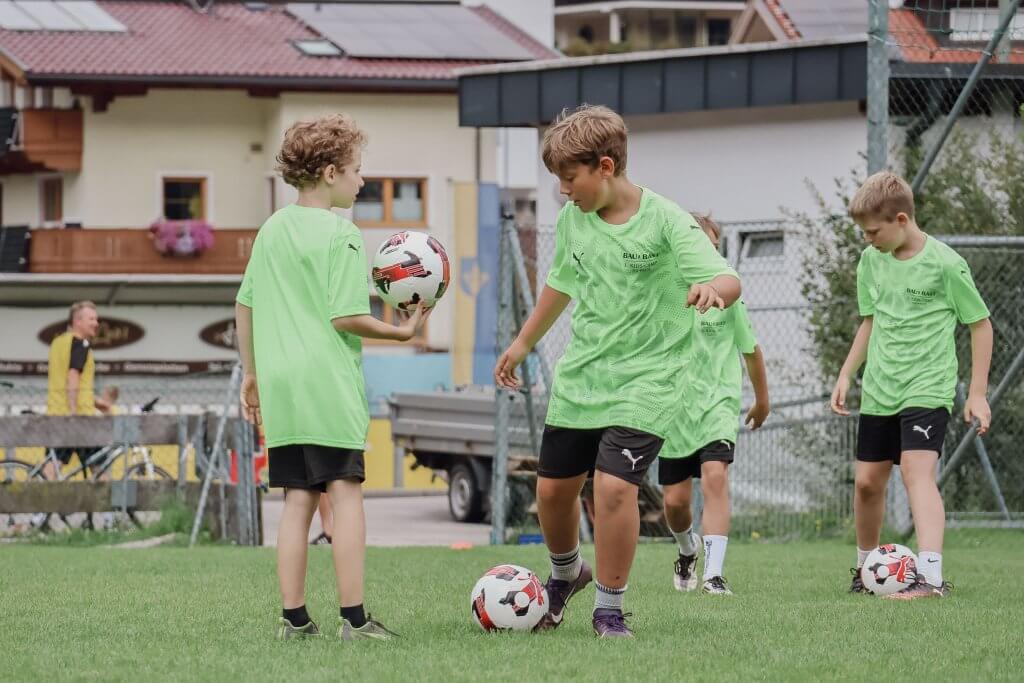 Kinder in grünen Trikots spielen Fußball auf einem Rasenplatz; Gebäude im Hintergrund.