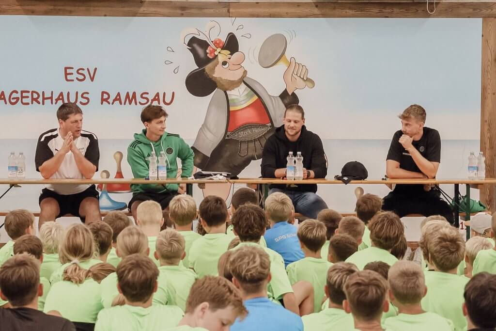 Vier Männer sitzen an einem Tisch vor einer Wand mit Zeichnung, umgeben von Kindern in grünen T-Shirts.