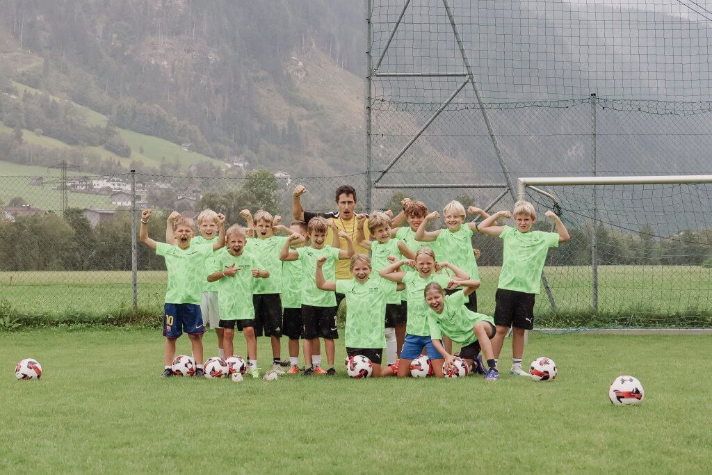 Kinder in grünen Trikots posieren mit einem Trainer auf einem Fußballplatz vor einem Tor.