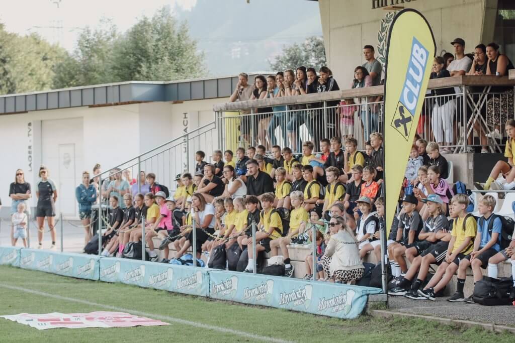 Kinder in Sportkleidung sitzen auf Tribünen bei einem Fußballturnier, umgeben von Zuschauern.