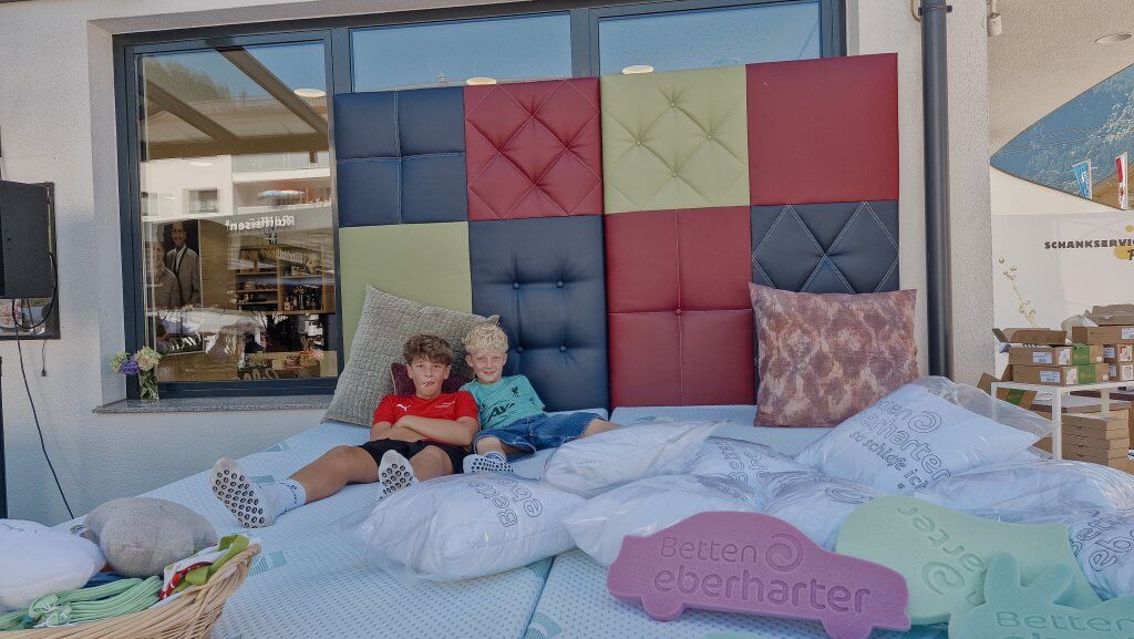 Zwei Jungen sitzen auf einem großen Bett mit buntem Kopfteil und dekorativen Kissen vor einem Geschäft.