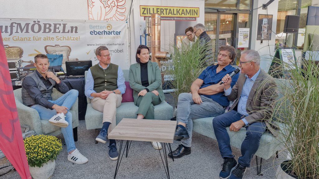 Fünf Personen sitzen auf Sofas bei einer Outdoor-Veranstaltung, umgeben von Werbebannern und Pflanzen.