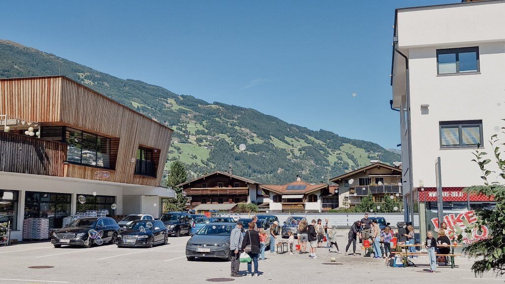 Menschen auf einem Parkplatz vor Geschäften in einer bergigen Gegend bei sonnigem Wetter.