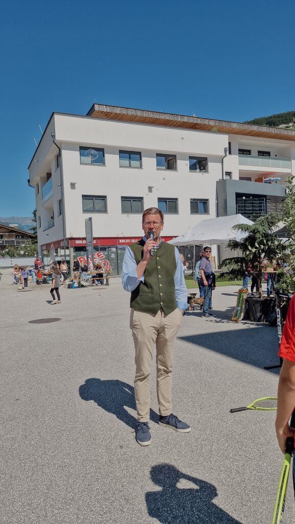 Mann mit Mikrofon spricht vor einem modernen Gebäude, umgeben von Menschen bei einer Outdoor-Veranstaltung.