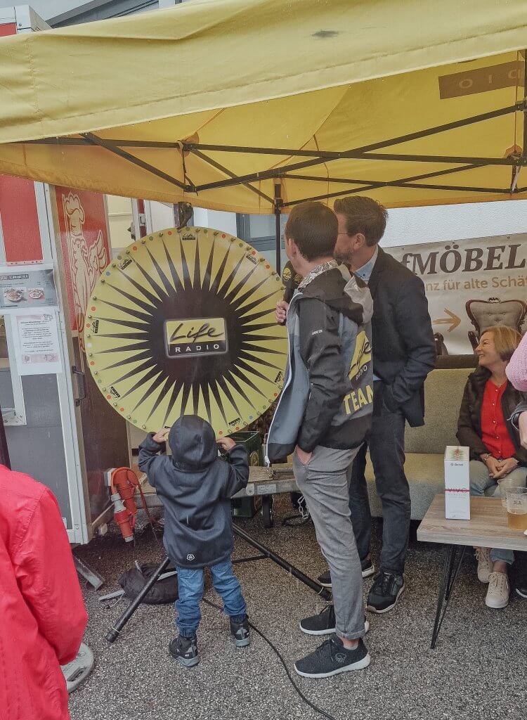 Kind dreht Glücksrad unter gelbem Pavillon, umgeben von Erwachsenen.