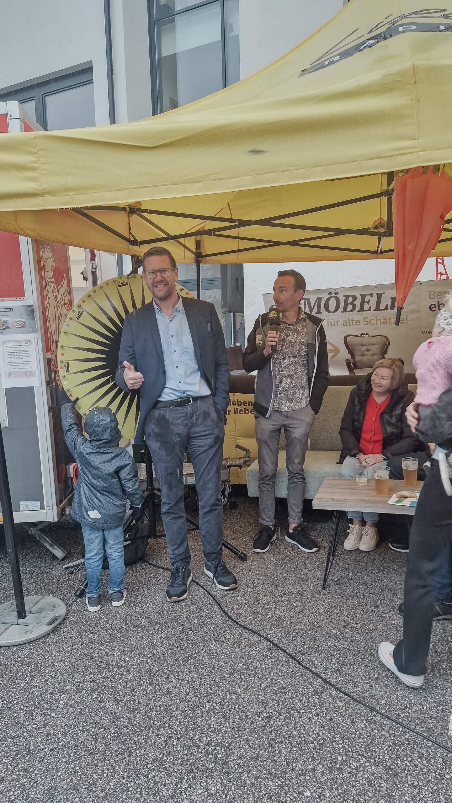 Mann in Anzug vor gelbem Zelt mit Glücksrad, Kind daneben, anderer Mann mit Mikrofon, Zuschauer auf Bank.