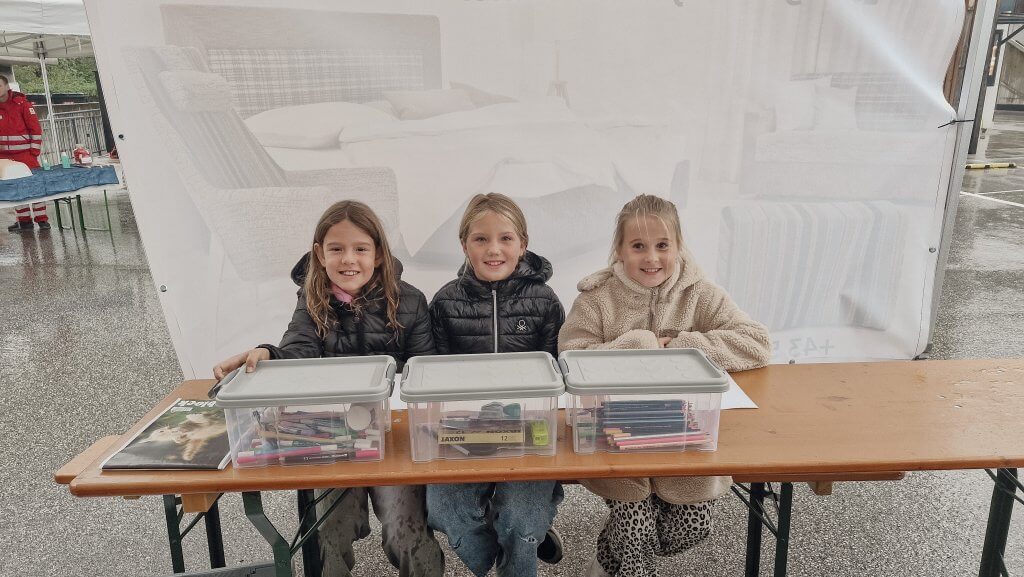 Drei Kinder sitzen an einem Tisch mit Bastelmaterial in Boxen, im Freien unter einem Zelt.